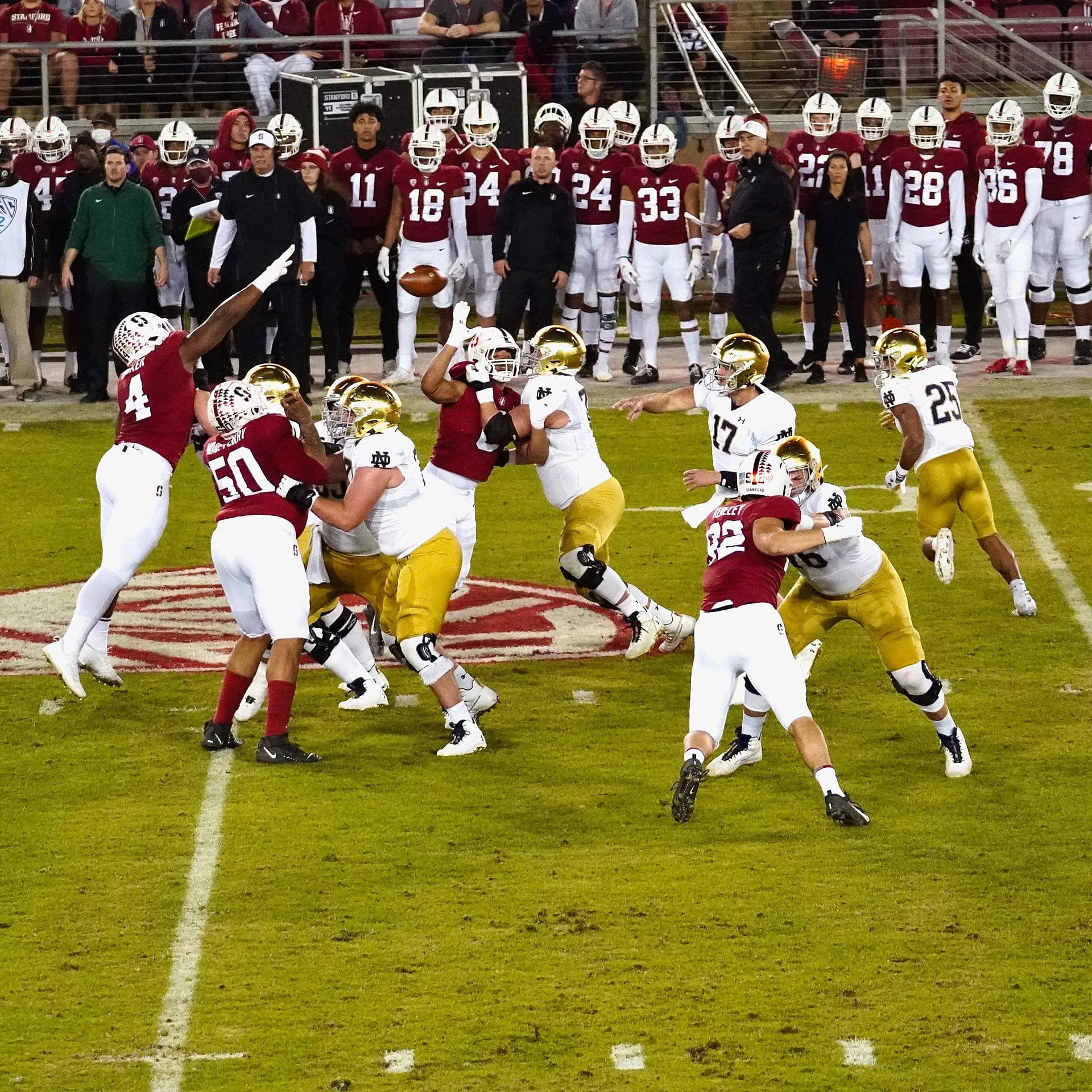 El mariscal de campo número 17 de Notre Dame lanza un pase mientras los defensores de Stanford, con uniformes color cardenal, avanzan y bloquean.