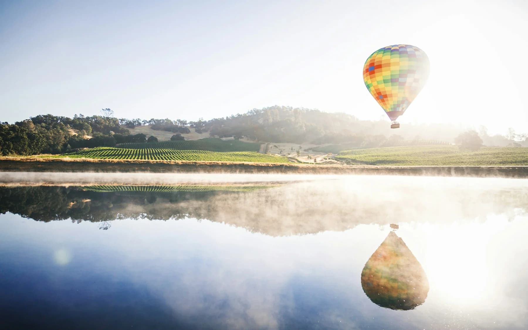 A colorful hot-air balloon above Napa vineyards reflected in still water and morning mist